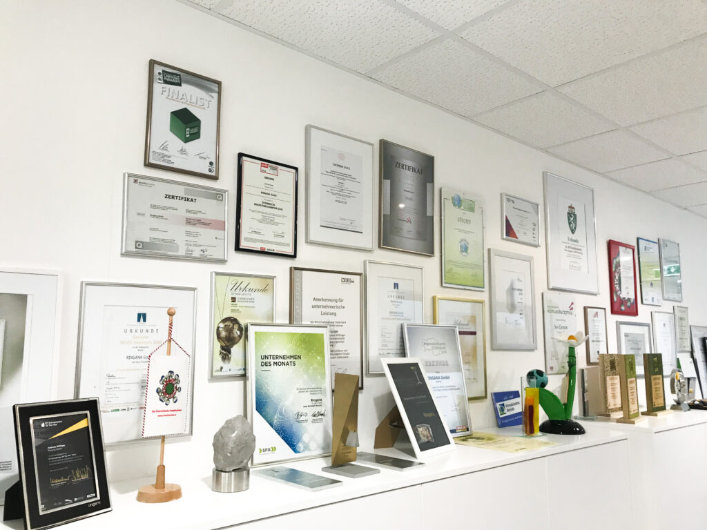 Wiele nagród, dyplomów i wyróżnień dla firmy RINGANA. Część z nich wisi na białej ścianie, a reszta jest ułożona na białym blacie